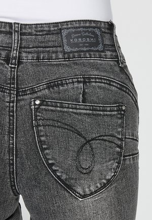 Fekete farmernadrág szív alakú hátsó zsebbel, márkás bőr címkével a derekán és kontrasztos varrási részletekkel.