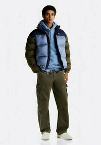 Tânăr îmbrăcat cu o jachetă puffer albastră, hanorac deschis la culoare, pantaloni cargo oliva și adidași albi, stând cu mâinile în buzunare.