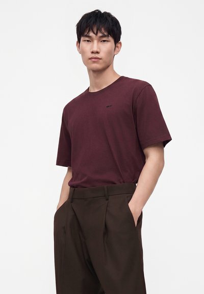 Jeune homme portant un t-shirt à manches courtes bordeaux et un pantalon marron foncé, les mains dans les poches, se tenant devant un fond blanc uni.