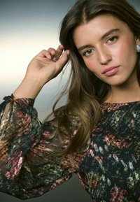 Sheer Bluse mit Blumenmuster in dunklen Tönen, die über Puffärmel und elastische Bündchen verfügt. Das Model spielt leicht mit den Haaren, während silberne Ohrringe sichtbar sind.