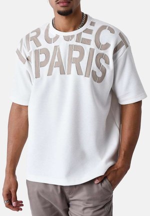 Camisa blanca de manga corta con tejido texturizado, que presenta "PROJECT PARIS" en grandes letras beige en la parte delantera. Cuello redondo y corte relajado.
