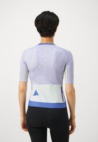 Korte ermer atletisk topp i lys lilla med mesh-detaljer, med hvit midjebånd og blå glidelåslomme. Teksturet stoff med logo.