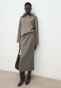 Sweter w kolorze khaki z suwakiem do ćwiartki i kołnierzem, połączony z dopasowaną ołówkową spódnicą. Noszony z brązowymi butami na obcasie i czarną torbą typu tote.