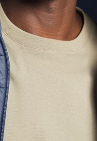 Beige lightweight cotton crew neck shirt with a soft texture, featuring a ribbed neckline. Partially visible underneath a blue zip-up jacket.