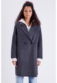 Abrigo de lana gris de doble botonadura con grandes solapas, bolsillos laterales y un corte relajado, superpuesto a un sudadera blanca con capucha y rayas.