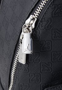 Zwart textuurmateriaal met een geëmbosseerd "23" patroon; zilveren rits hardware met een Jumpman-logo detail; slank en modern ontwerp.
