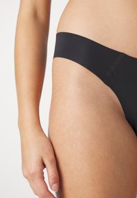String noir en tissu lisse et extensible. La ceinture est large et plate, et le design est minimal avec aucune couture visible.