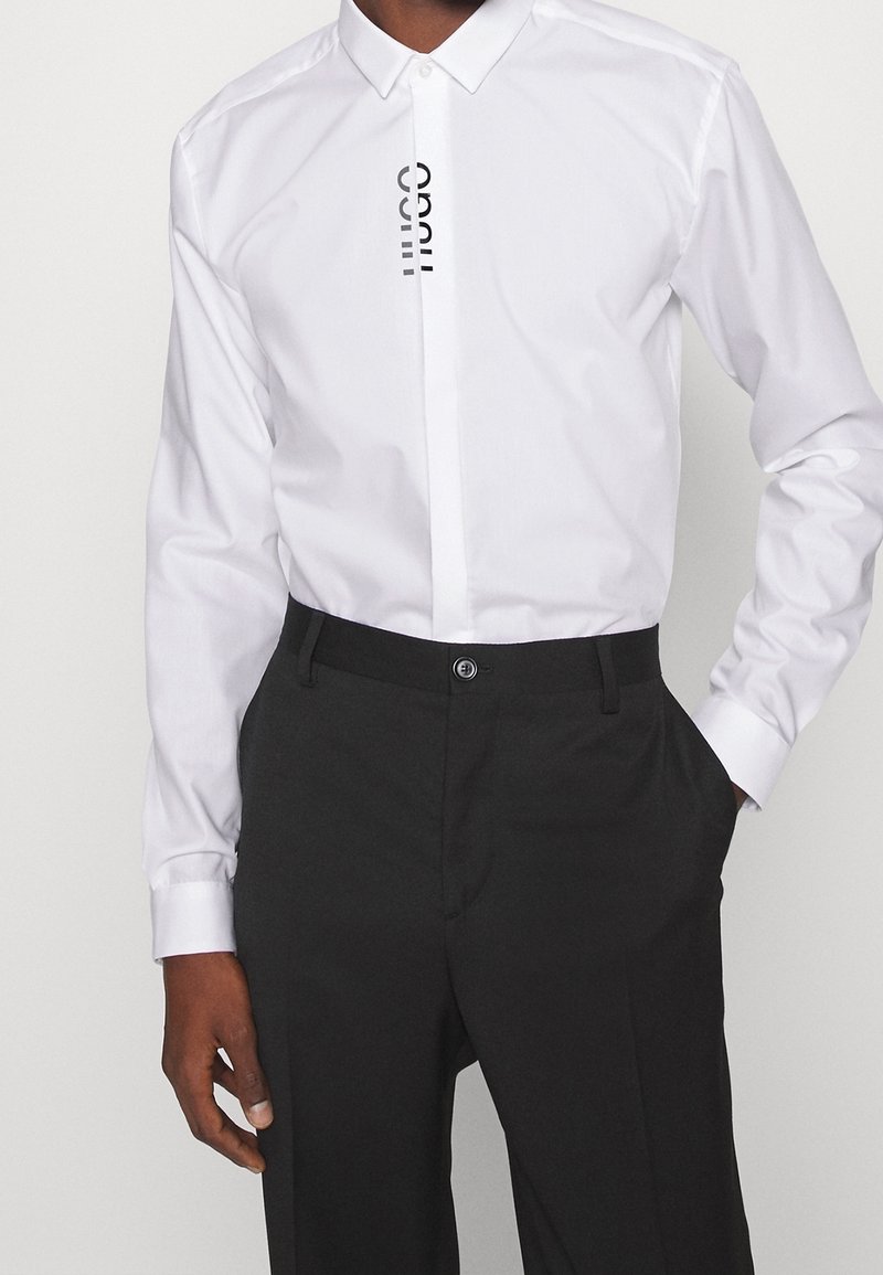 Witte blouse met knopen, featuring verticale zwarte logo op de voorkant, gecombineerd met donkere broek. Gladde stof met klassieke kraag en lange mouwen.