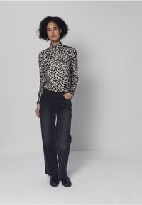 Leopardmönstrad långärmad topp i mjukt tyg med hög krage, kombinerad med lösa svarta jeans. Modellen står med händerna i fickorna mot en neutral bakgrund.