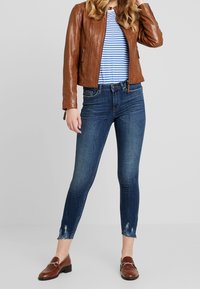 Hnědá kožená bunda, modro-bílá pruhovaná košile, tmavě modré skinny džíny s třepenými lemy, hnědé mokasíny, celkově neformální outfit.