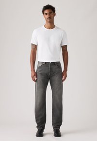 Άνδρας που στέκεται φορώντας ένα απλό λευκό t-shirt, ξεθωριασμένα μαύρα τζιν, μαύρα παπούτσια, με τατουάζ και στα δύο αντιβράχια, μπροστά σε απλό φόντο.