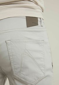 Pantalon gris clair avec une texture lisse, présentant un patch rectangulaire portant l'étiquette "CHASINI" et une couture distinctive en forme de triangle sur la poche.