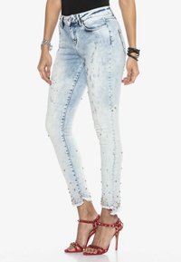 Cienkie dżinsy z jasnego denimu, z postarzanymi detalami i ozdobnymi brzegami zdobionymi koralikami. Noszone z czerwonymi, cekinowymi sandałami na wysokim obcasie.