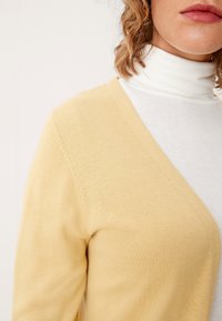 Dziany żółty kardigan z dekoltem w serek, prezentujący miękką fakturę. Nosi się go na białym golfie, podkreślając kontrastujące warstwy.