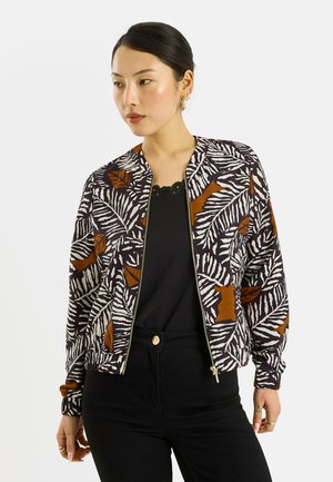 Mujer con una camiseta y pantalones negros y una chaqueta con cremallera con estampado de hojas en negro, blanco y marrón, mirando hacia su izquierda.