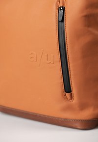 Tasche aus braunem Leder mit strukturierter Oberfläche, ausgestattet mit einem schwarzen Reißverschluss und geprägtem Branding an der Seite. Kompakte rechteckige Form.