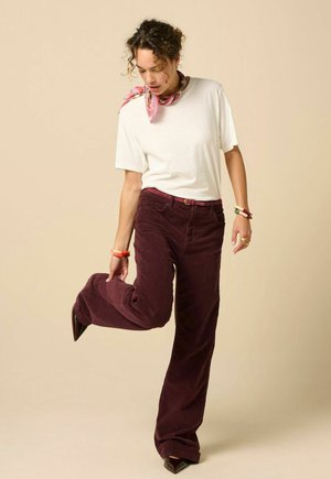 Femme portant un t-shirt blanc, un pantalon large bordeaux et une écharpe rose ajuste sa chaussure, debout devant un fond beige.