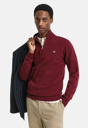 Burgunderfarbener Pullover mit Reißverschluss aus weichem Stoff, ausgestattet mit gerippten Bündchen und Saum, kombiniert mit einem navyblauen Blazer mit Streifen, der über die Schulter gelegt ist.