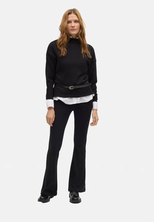 Suéter negro de canalé con un cuello alto, llevado sobre una camisa blanca de botones. Unos pantalones negros ajustados y unos zapatos negros brillantes completan el look.