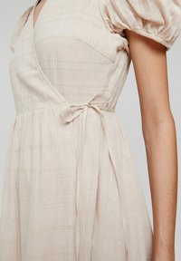 Robe portefeuille en tissu beige clair à rayures texturées, manches courtes bouffantes et ceinture à la taille. Elle présente un col en V et une silhouette fluide.