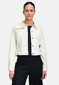 Veste en denim blanche avec fermeture à boutons, deux poches poitrine, texte brodé sur le côté gauche et accent de palmier sur la manche droite.