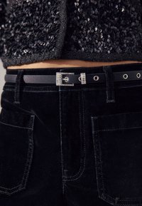 Svarta sammetsbyxor med hög midja och ett brett bälte med dekorativ spänne av strass, metalliska öljetter och dubbla fickor fram.