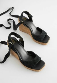 Sandali con zeppa nera che presentano una tomaia in tessuto intrecciato, punta quadrata e una piattaforma in juta naturale testurizzata. Cinghie regolabili alla caviglia con dettagli in oro.