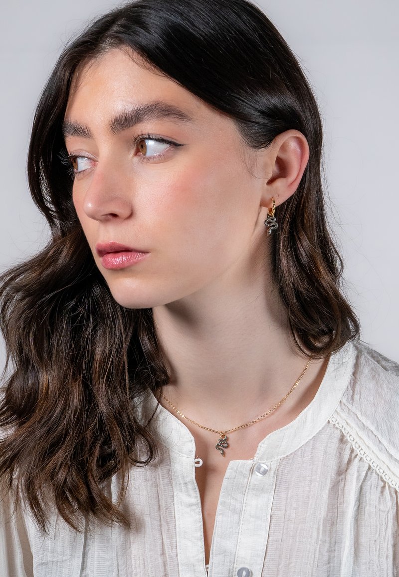 Guldörhängen med en liten ormdesign, tillsammans med ett fint guldhalsband med en liknande berlock, buren av en modell.