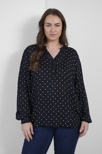 Blouse noire à manches longues avec des pois blancs, présentant un décolleté en V, une patte de boutonnage et une coupe décontractée, assortie à un jean bleu.