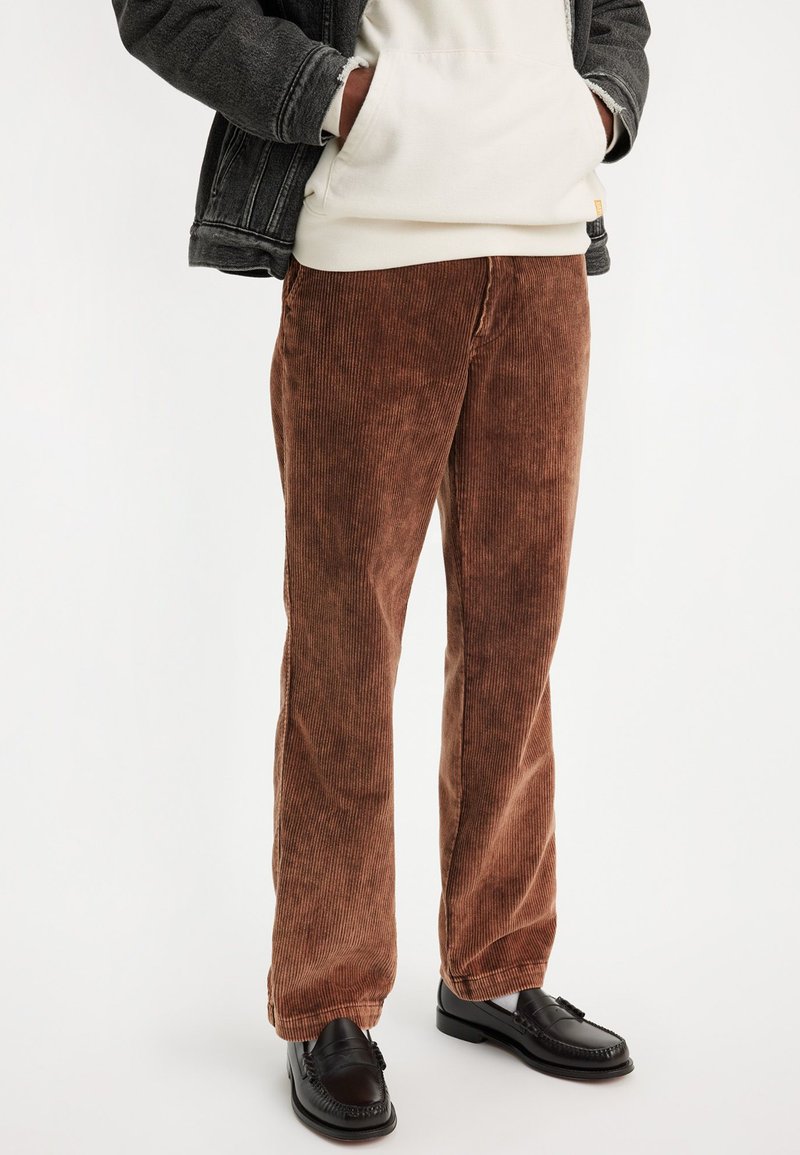 Braune Cordhosen mit vertikalen Streifen, geradem Schnitt und einer weichen Textur. Kombiniert mit schwarzen Slippern und einer passenden Jacke.