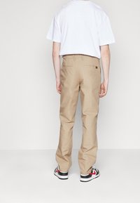 Pantalones de algodón beige con corte relajado, que cuentan con bolsillos traseros y una cinturilla. Una camiseta blanca básica completa el conjunto.