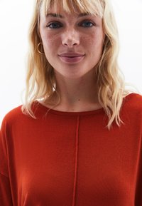 Roter Strickpullover mit rundem Ausschnitt und einer zentralen Naht. Das Material hat eine weiche Textur. Das Model hat hellblonde Haare.