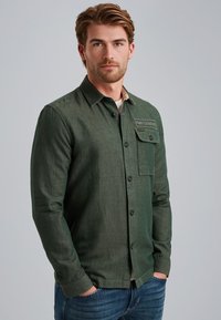 Chemise à manches longues vert foncé avec une poche poitrine, boutonnière à l'avant et un col beige contrastant. Tissu texturé avec un motif subtil.