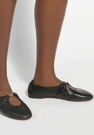Femme portant des chaussures plates en cuir noir avec une petite boucle décorative dorée sur le dessus, sur fond blanc.