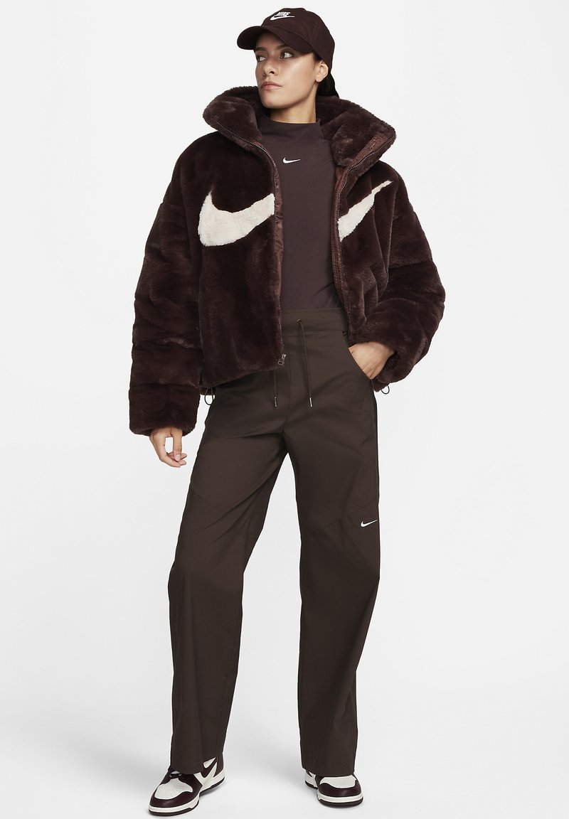 Giacca in finta pelliccia marrone con logo swoosh crema, abbinata a pantaloni marrone scuro e un berretto abbinato con un logo. Sneakers con dettagli bianchi.
