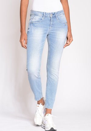 Personne portant un jean slim bleu clair et des baskets blanches, debout devant un fond clair uni.