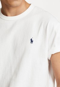 Vitt bomullsis t-shirt med upprullade ärmar, med en liten marinblå polo-spelare logotyp på bröstet. Slät, lätt textur.