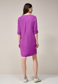 Lila linnklänning med trekvartsärmar, upprullade manschetter och en bakfålla, med en rak silhuett och sidoklyftor.