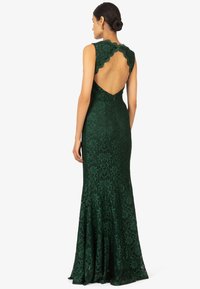 Grünes Spitzenkleid mit gewelltem Ausschnitt und offener Rückseite. Enganliegendes Oberteil, ausgestellteter Rock, florales Muster, verdezipfelter Reißverschluss an der Rückseite. Lange Länge.