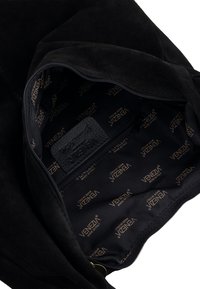 Wnętrze czarnej zamszowej torby posiada podszewkę z logo oraz skórzaną metkę. Widoczna jest kieszeń na zamek, ukazująca design torby.