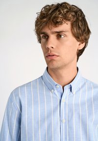 Camicia azzurra chiara con righe verticali bianche, colletto appuntito e bottoni piccoli; il tessuto sembra morbido e con una texture.