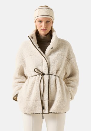 Frau trägt eine cremefarbene Fleecejacke mit Taillengürtel, beigen Rollkragen, passendes gestricktes Stirnband und helle Hosen, blickt nach vorne.
