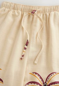 Pantalon en tissu beige avec taille élastique et cordon à nouer présentant des motifs décoratifs violets et jaunes près de l'ourlet.