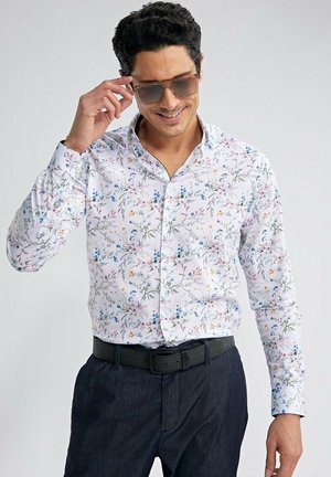 Homme portant des lunettes de soleil, une chemise blanche avec un imprimé floral coloré, un pantalon sombre et une ceinture noire, ajustant ses lunettes de soleil avec sa main droite.