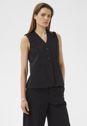 Femeie stând cu fața înainte, purtând o vestă neagră fără mâneci, cu nasturi și două buzunare frontale, precum și pantaloni negri asortați, pe un fundal simplu.