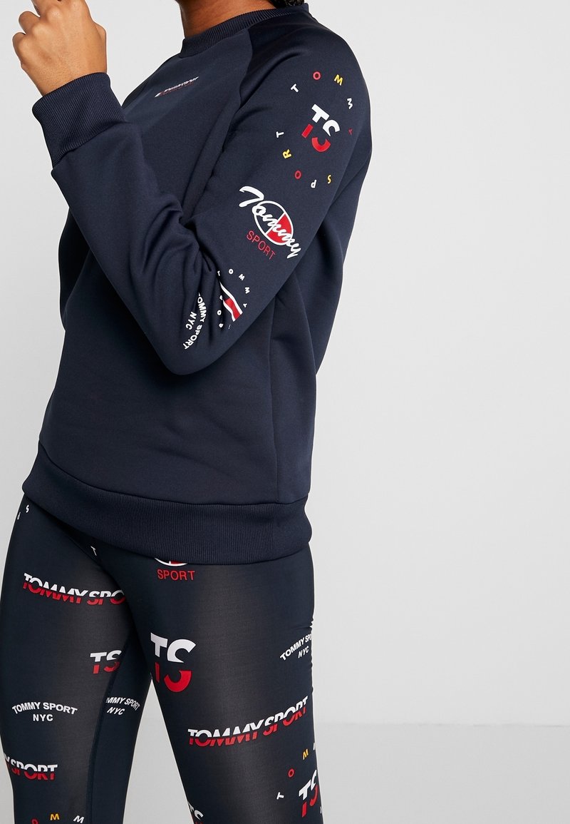 Granatowy sweter z ribbami przy mankietach i dolnym końcu, z kolorowymi wzorami logo na rękawach. W zestawie z legginsami w podobne kolory.