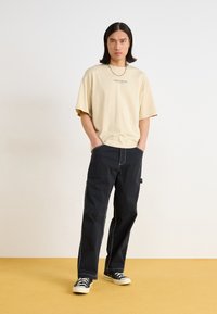 Beige oversized t-skjorte med trykket logo, kombinert med svarte vide bukser med sidelommer og hvit søm. Svarte joggesko fullfører antrekket.
