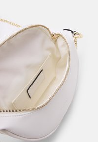 Bílá veganská kožená taška se zipem v zlaté barvě, vnitřní kapsou na karty a béžovou textilní podšívkou. Zakulacený tvar s minimalistickým designem.