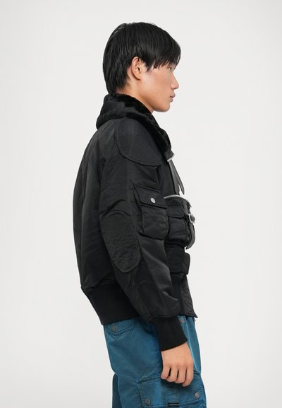 Cazadora negra tipo bomber con un cuello de piel sintética, parches texturizados en los hombros, detalles con cremallera y múltiples bolsillos frontales, combinada con pantalones azules.