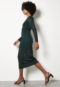 Un vestido de encaje verde esmeralda con mangas largas, dobladillo con forma de ondas y una silueta ajustada. Combinado con sandalias negras de tiras. Detalles de tejido texturizado visibles.